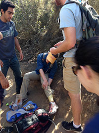 man sitting down on the ground clutching his leg while two emergency team personnel walk toward him 